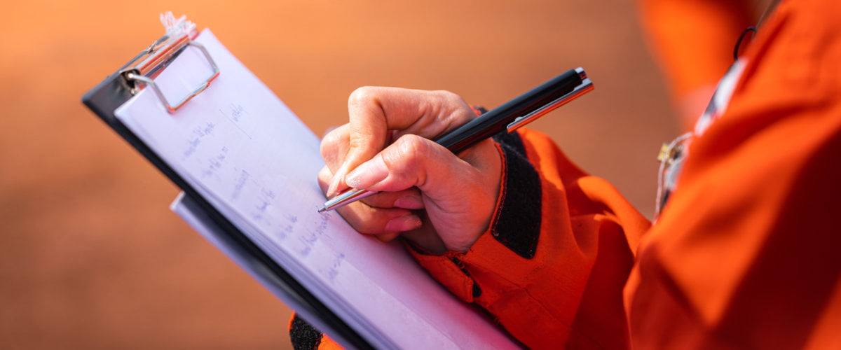 Close up of Health and Safety Officer writing notes on a paper checklist during the audit and inspection of an oil field operation.