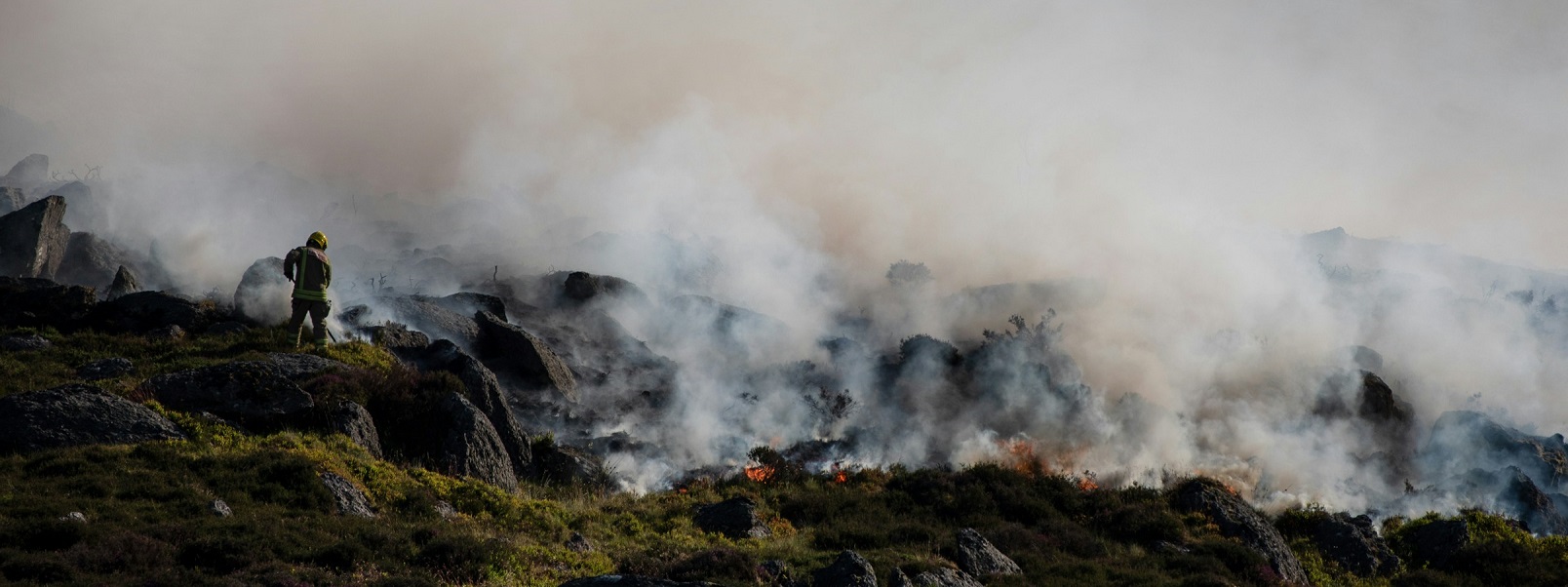 Photograph of a wildfires producing lots of smoke on Y Fron in Wales with a firefighter working to control the fire.