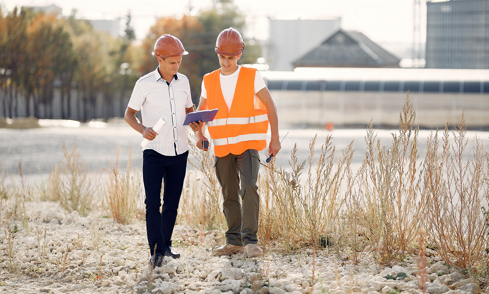 Photograph of two workers wearing hard hats, one wearing a high visibility jacket, operating a buddy sytem whilst walking around the outside of a rurally located factory.
